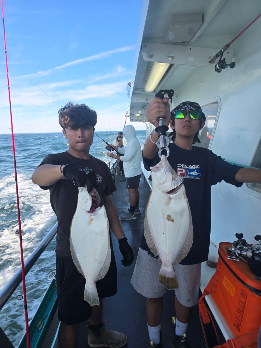 Two people on a boat holding flatfish, with ocean in the background.
