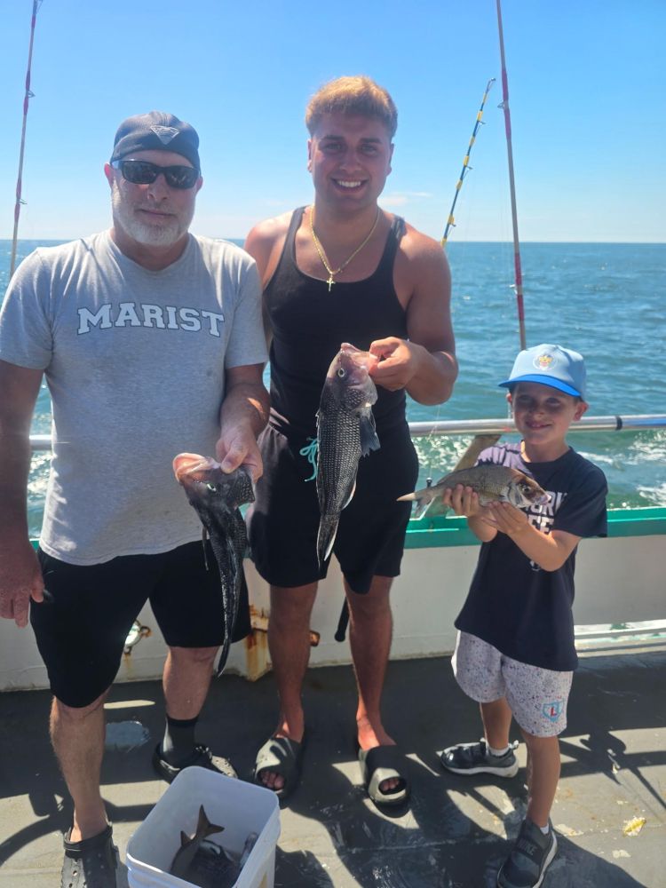 Three people on a boat holding fish, with fishing rods and ocean in the background.