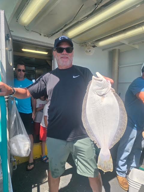 Man in sunglasses holding a large fish on a boat with people in the background.