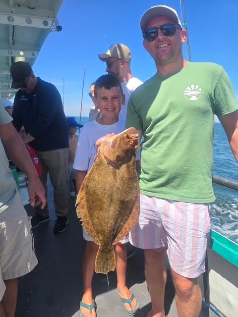 Man and boy on a boat holding a large flatfish under a sunny sky.