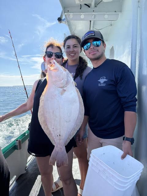 Three people on a boat holding a large flatfish, with ocean in the background.