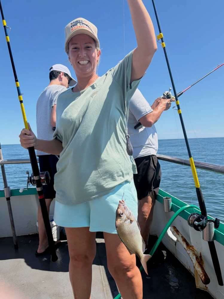 Person on a boat holding a fishing rod with a fish, smiling under a clear blue sky.