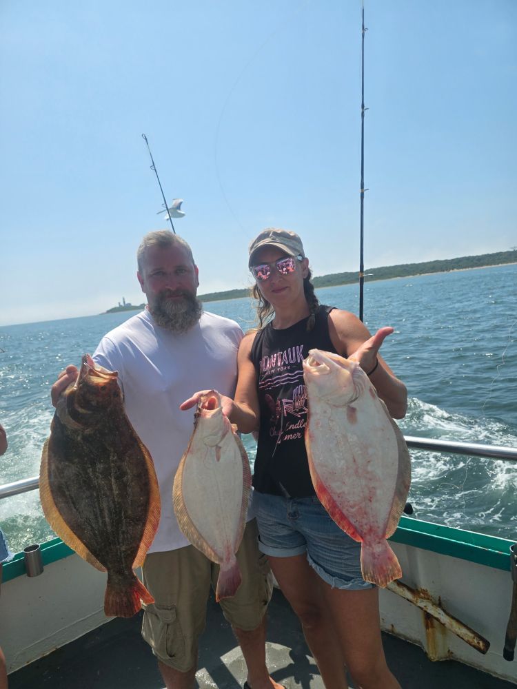 Two people on a boat holding three large fish with ocean in the background.