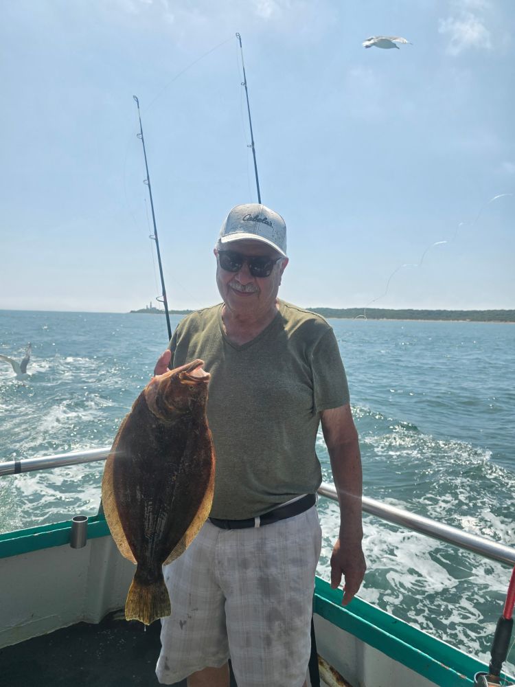 Man holding a large fish on a boat with ocean backdrop and fishing rods.
