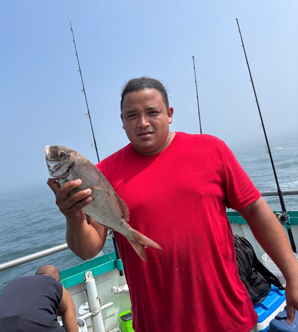 Man in red shirt holding a fish on a boat with fishing rods in the background.