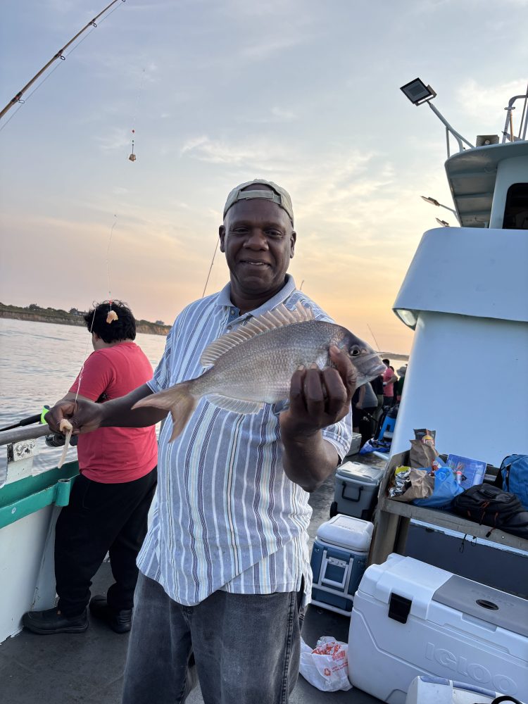 Man holding a fish on a boat while smiling at sunset.