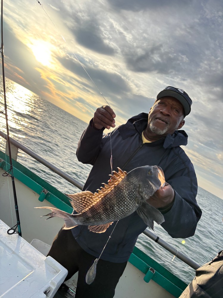 Man on boat holding a fish at sunset, calm sea in background.