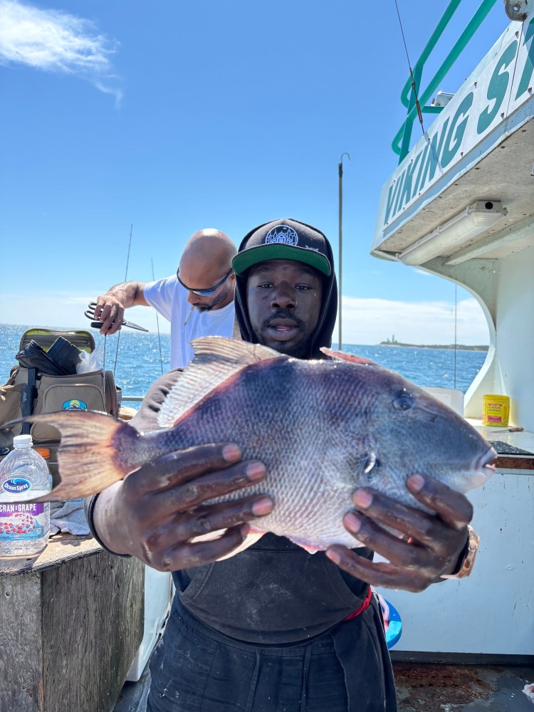 Man holding a fish on a boat, another person adjusts their gear in the background.