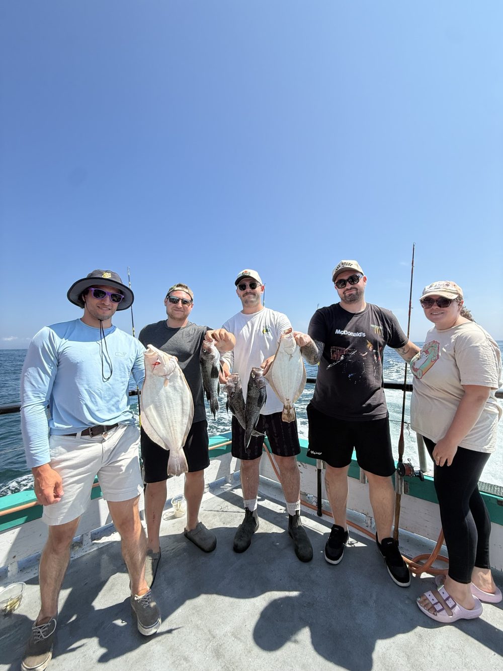 Six people standing on a boat with fishing rods and caught fish under clear sky.