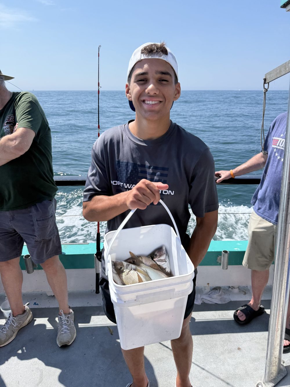 Person holding a white bucket with fish on a boat, smiling, with the ocean in the background.