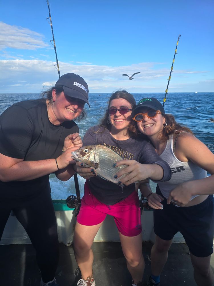 Three people on a boat holding a fish, smiling, with a seagull in the background.