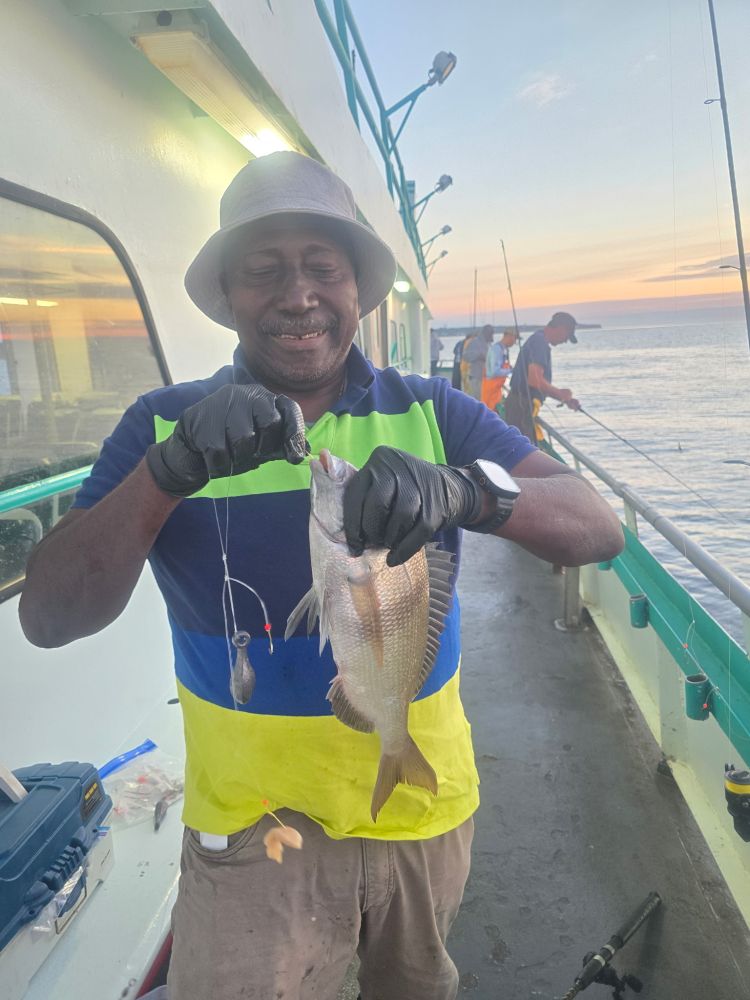 Man on a boat holding a fish, smiling at sunset with fishing gear.