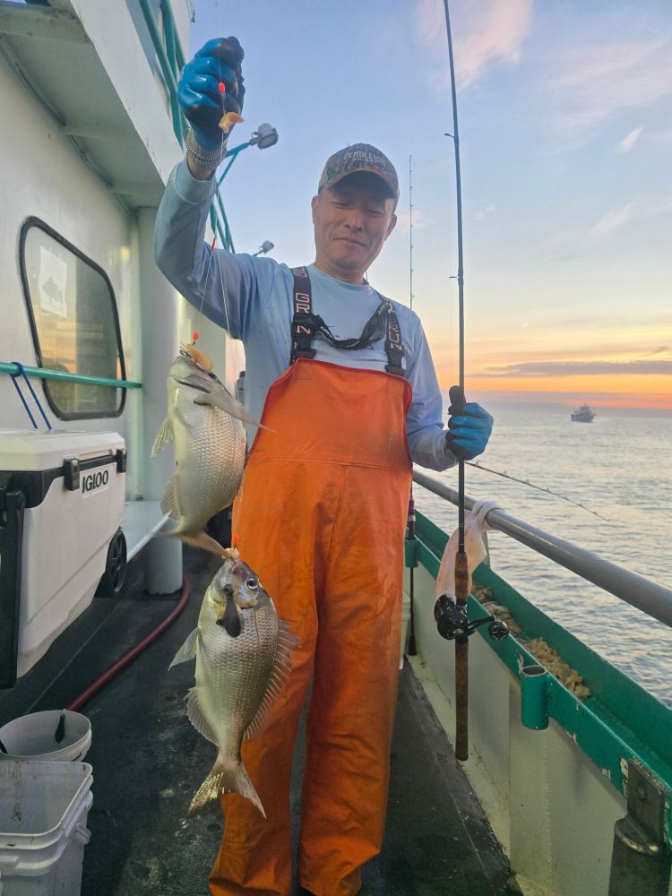 Man in orange overalls holds fishing rod with three fish on boat at sunset.