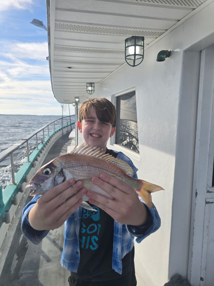 Boy smiling, holding a fish on a boat deck with ocean in the background.