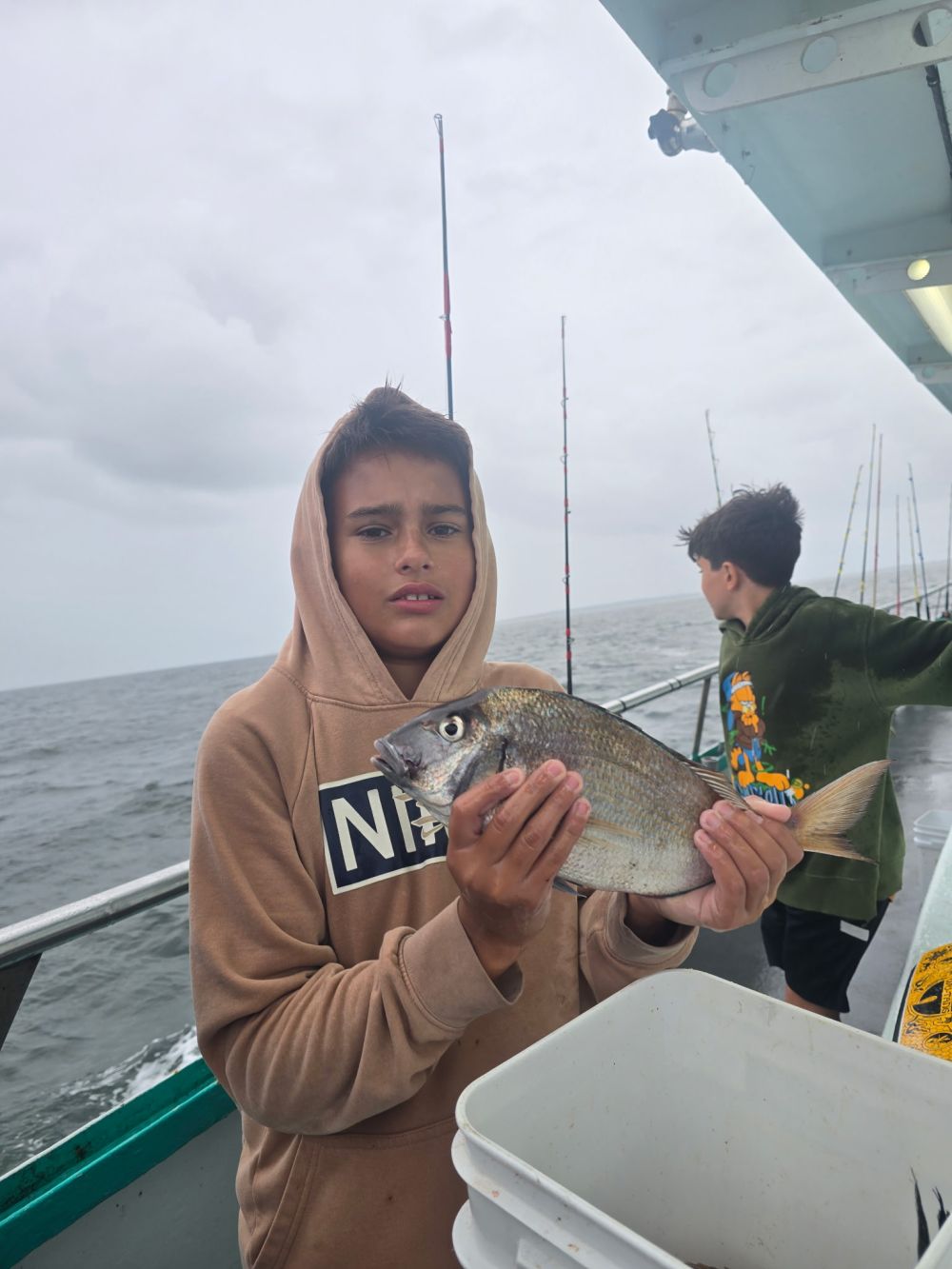 Boy holding a fish on a boat with another person and fishing rods in the background.
