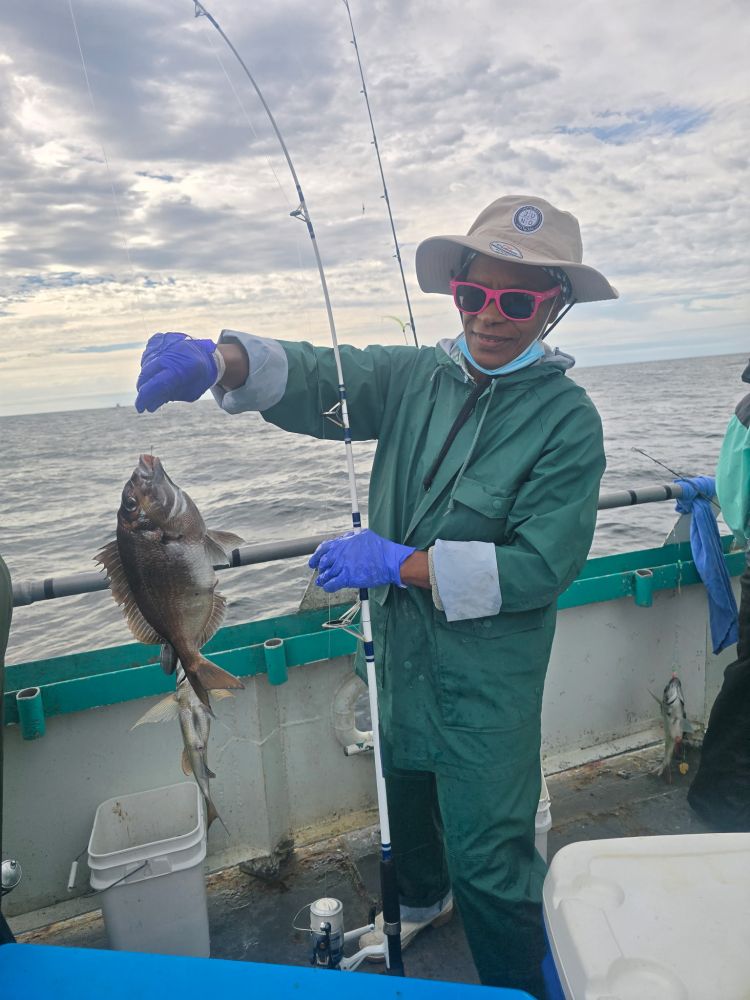 Person on a boat holding a fish, wearing a hat, sunglasses, and gloves, with ocean in the background.