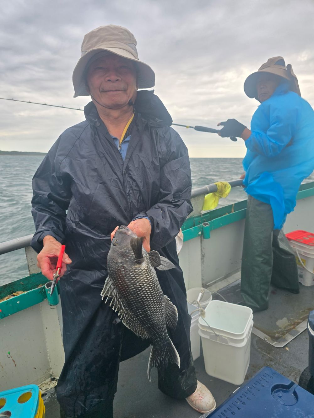 Two people fishing on a boat; one man holds a fish and a tool.