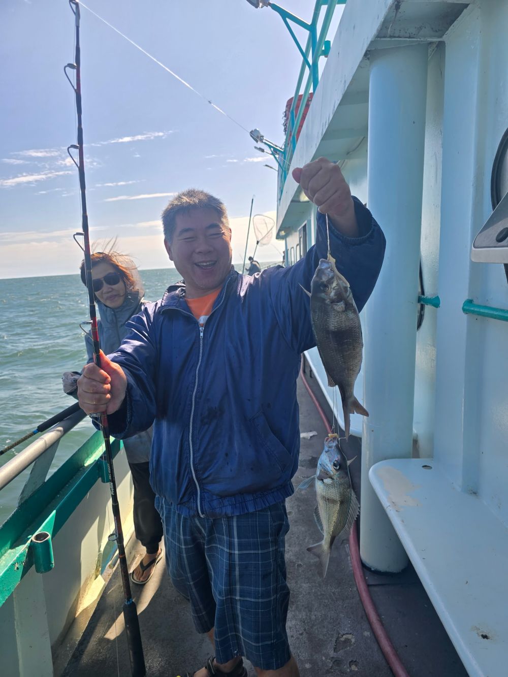Two people on a boat hold fishing rods and a caught fish with a net in the background.