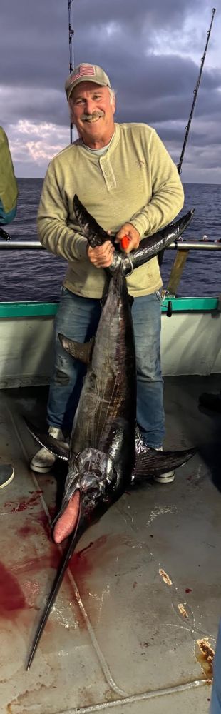 Man holding large swordfish on a boat at sea, with a cloudy sky in the background.