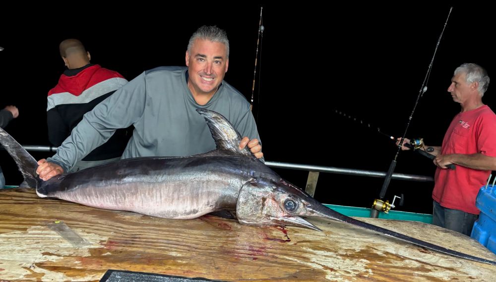 Man smiling with a large swordfish on a boat at night.