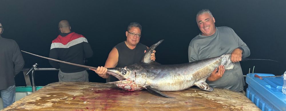 Two men on a boat hold a large swordfish on a wooden table at night.