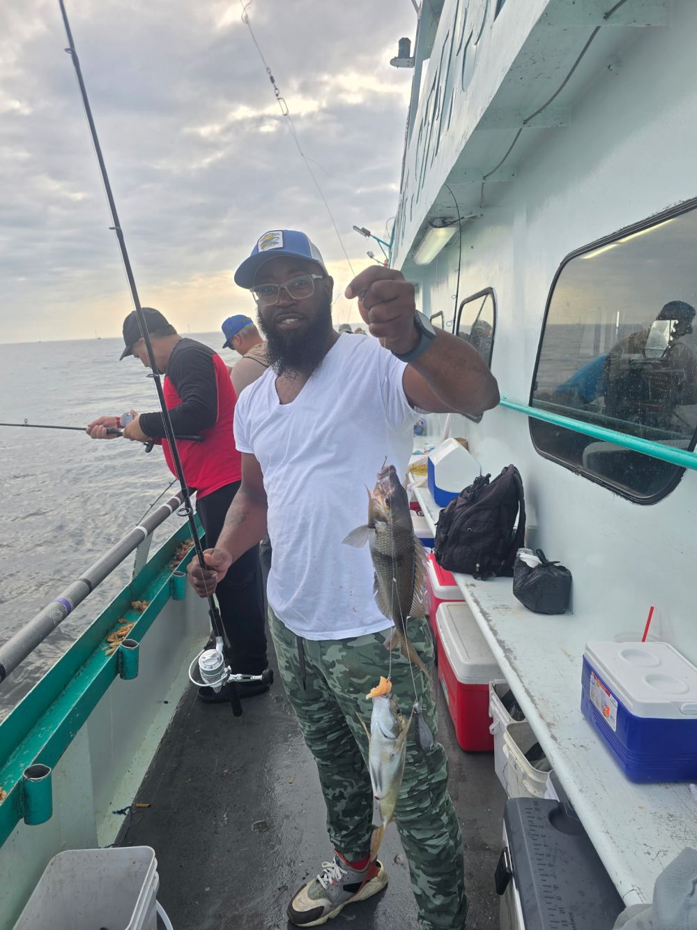 Man on boat holding two fish with fishing rod; cloudy sky.