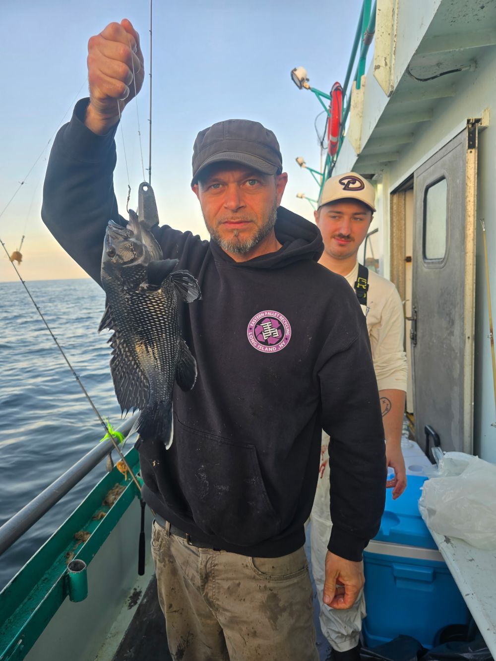 Two men on a boat holding a caught fish, with ocean seen in background.