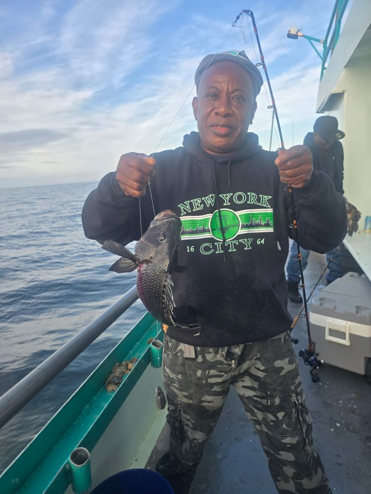 Man holding a fish on a fishing boat, wearing a New York City hoodie.