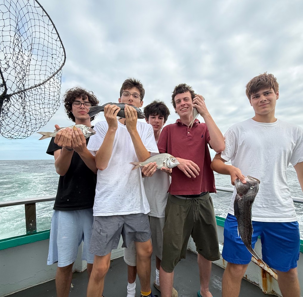 Five people on a boat holding fish, with ocean and cloudy sky in background.