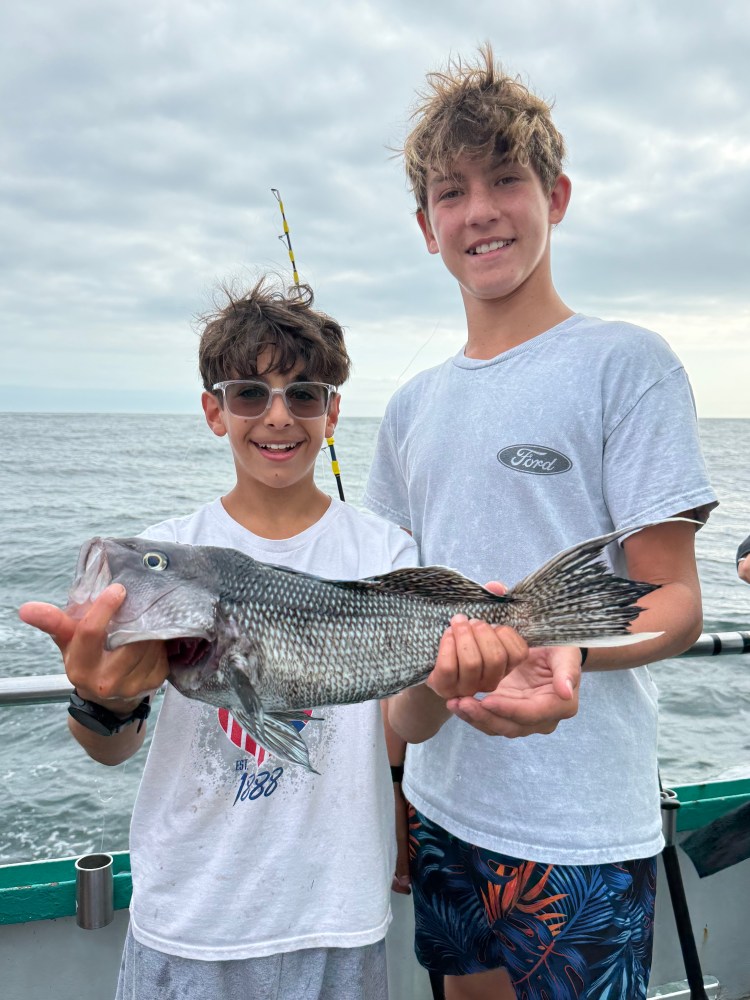 Two boys holding a large fish on a boat, with cloudy sky in the background.