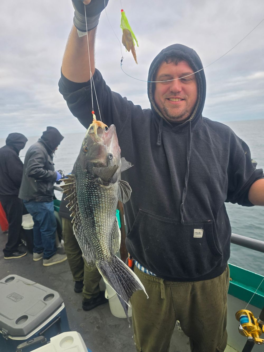 Man in hoodie smiles, holding a freshly caught fish on a boat.