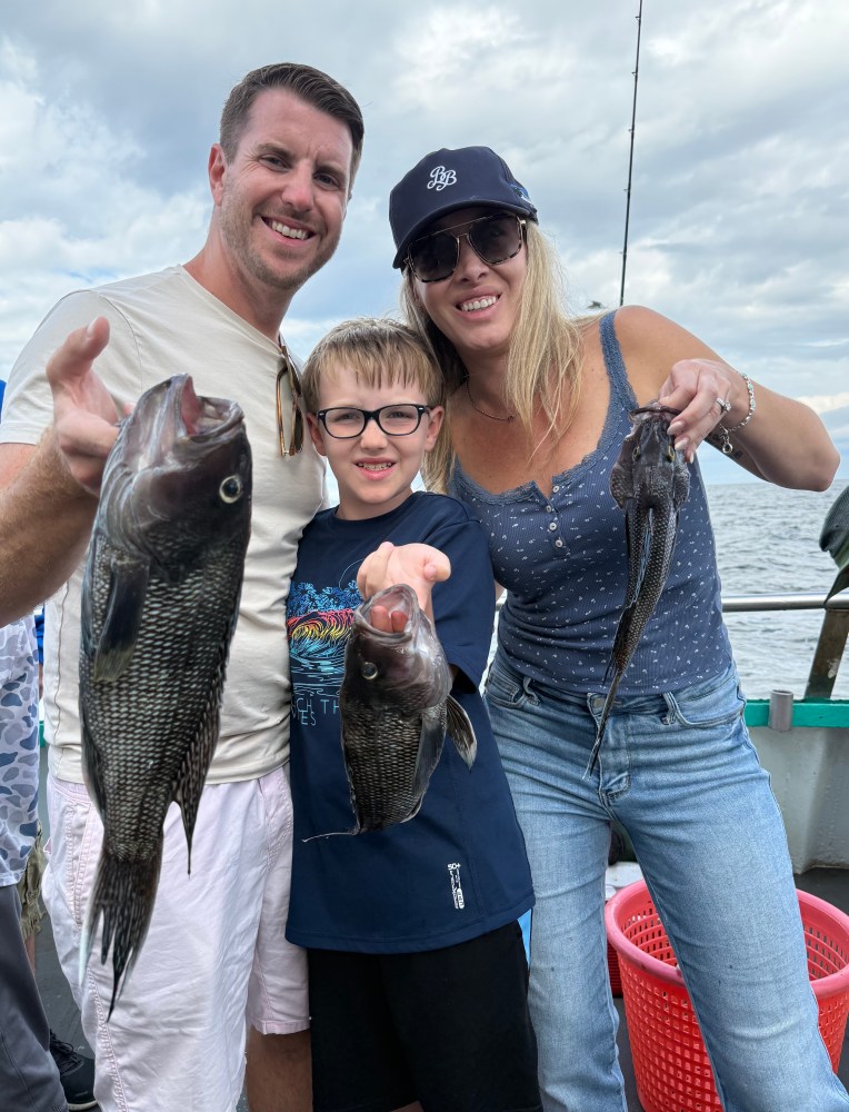 Three people smiling and holding two large fish on a boat.