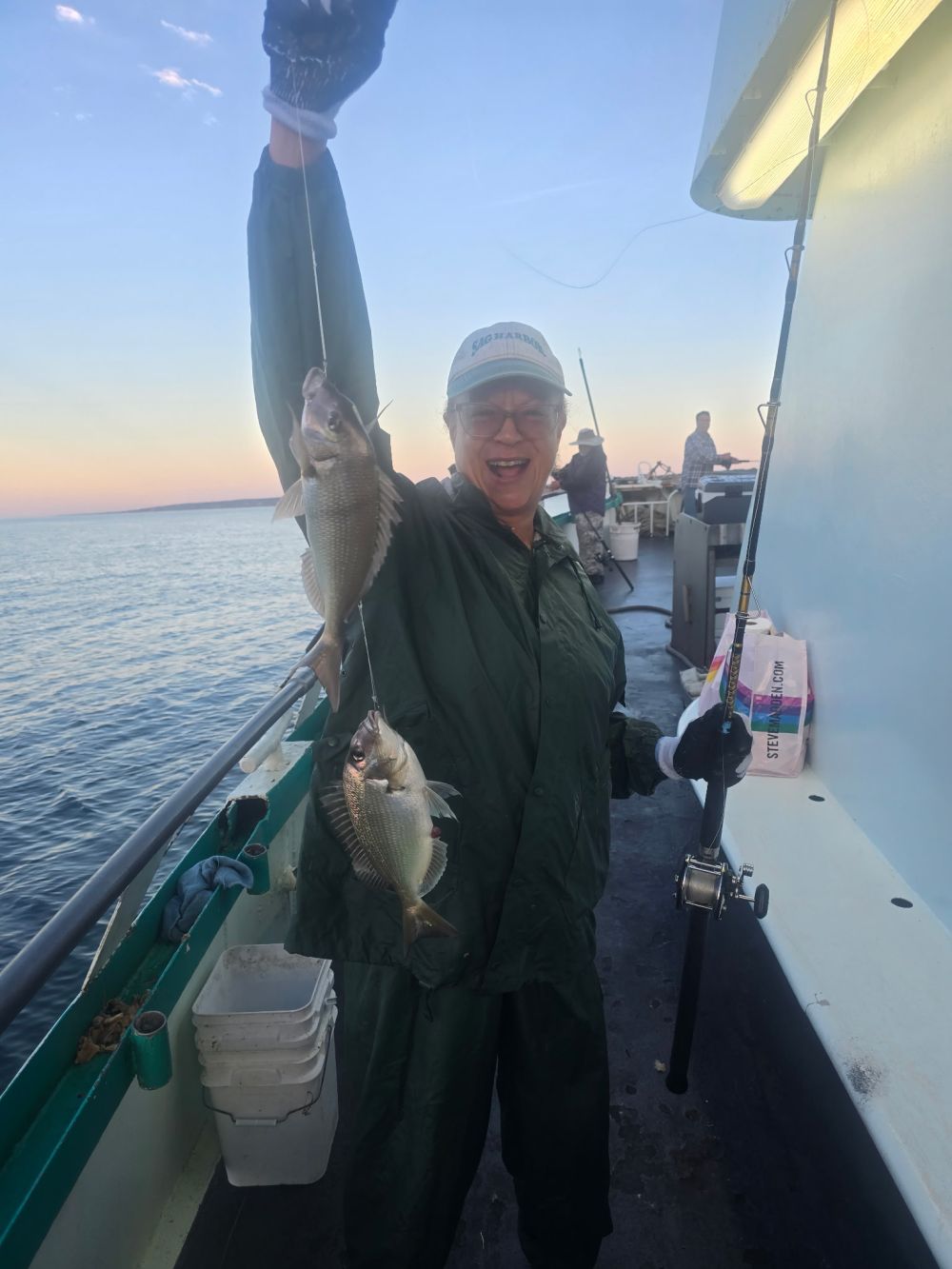 Person on boat holding multiple caught fish with a fishing rod at sunset.