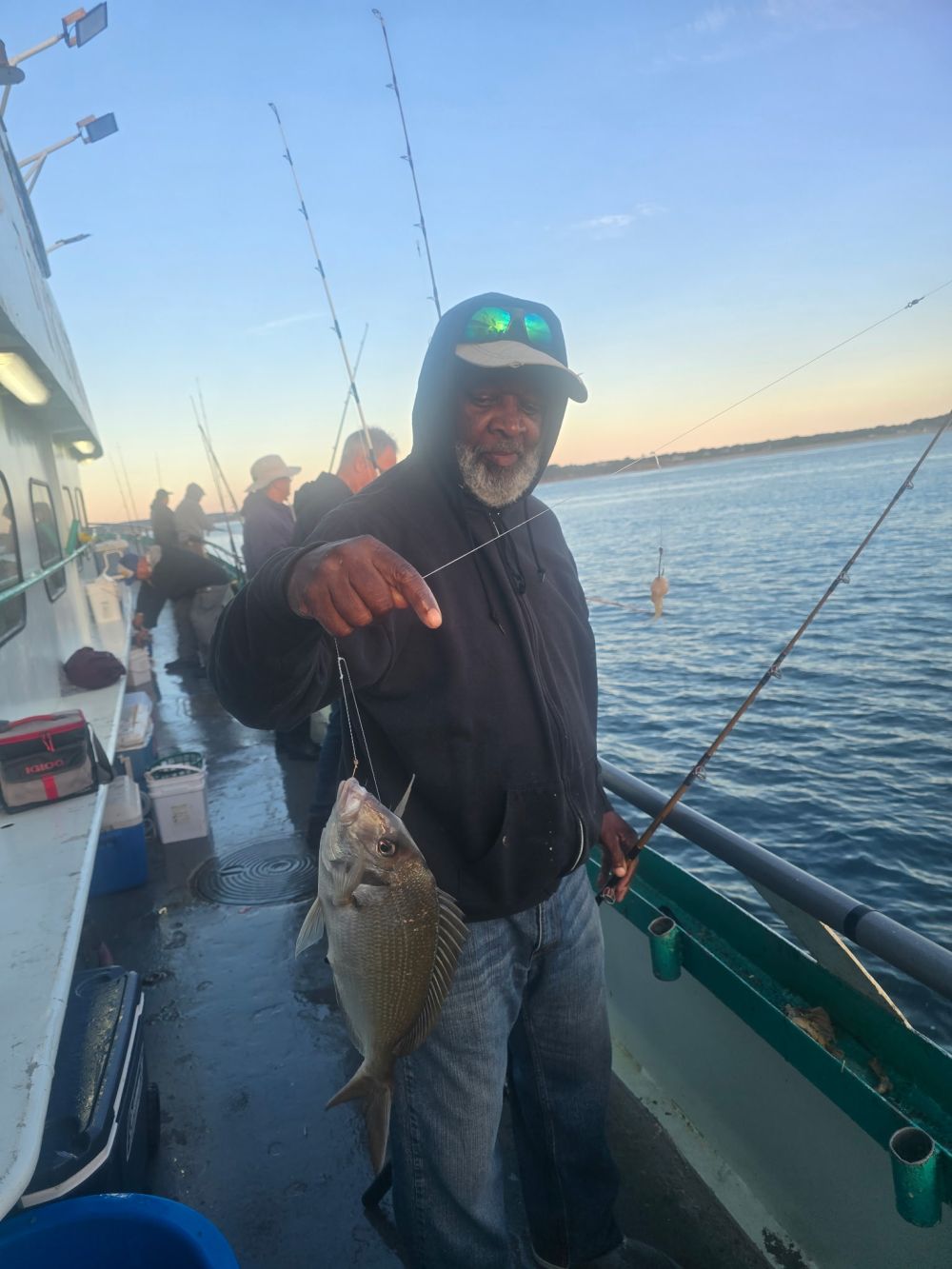 Man on boat holding a fish with fishing rods and water in background.