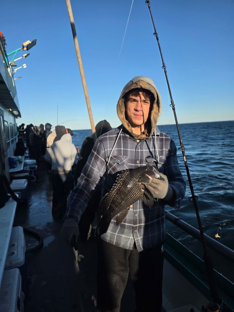 Person on boat holding fish, wearing hooded jacket, others in background, ocean view.