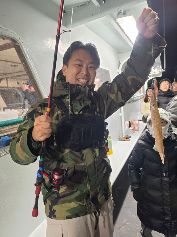 Person in camo jacket proudly holding a fishing rod with a small fish on a boat.