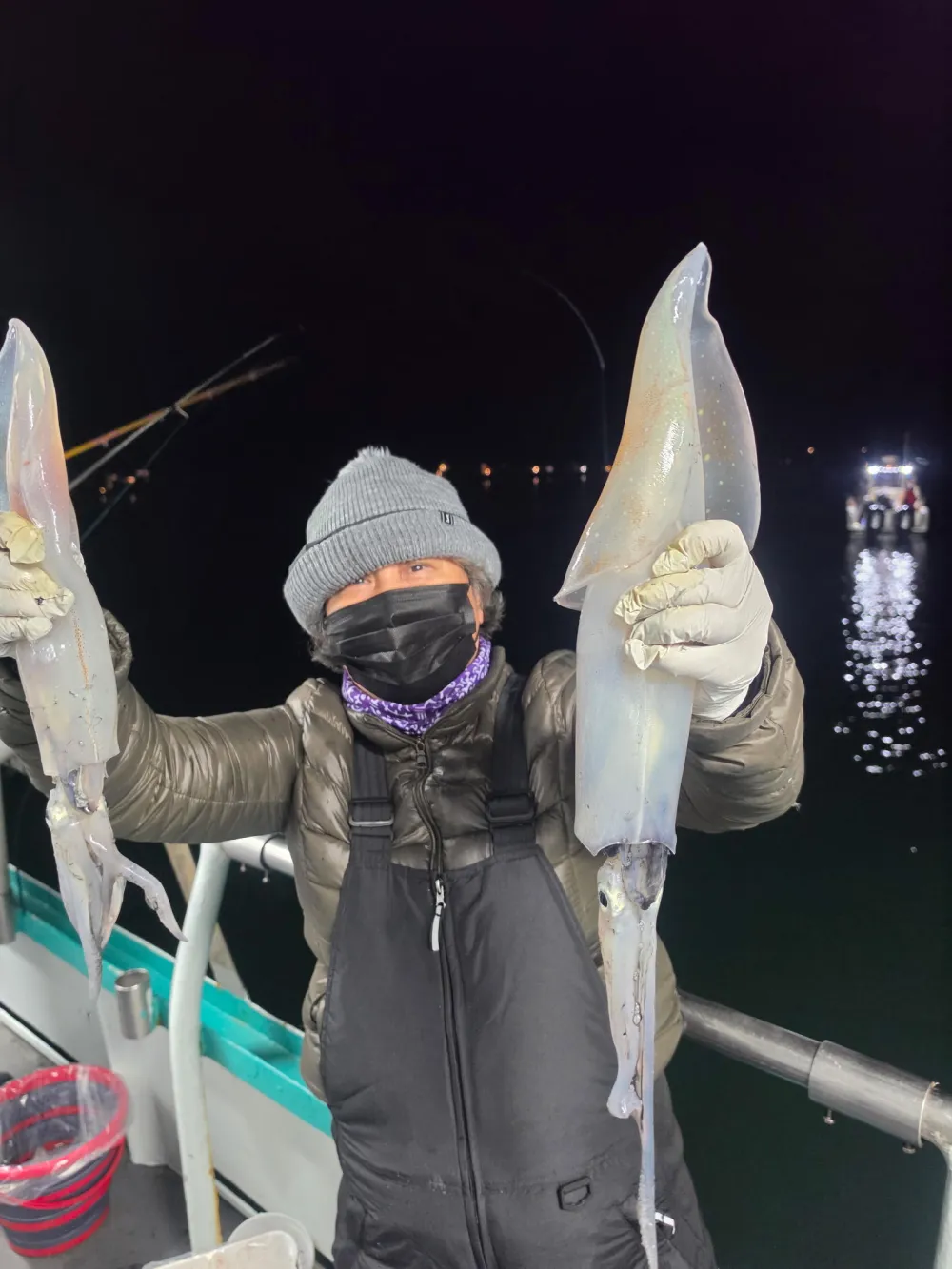 Person in winter clothing holds two large squids on a boat at night.