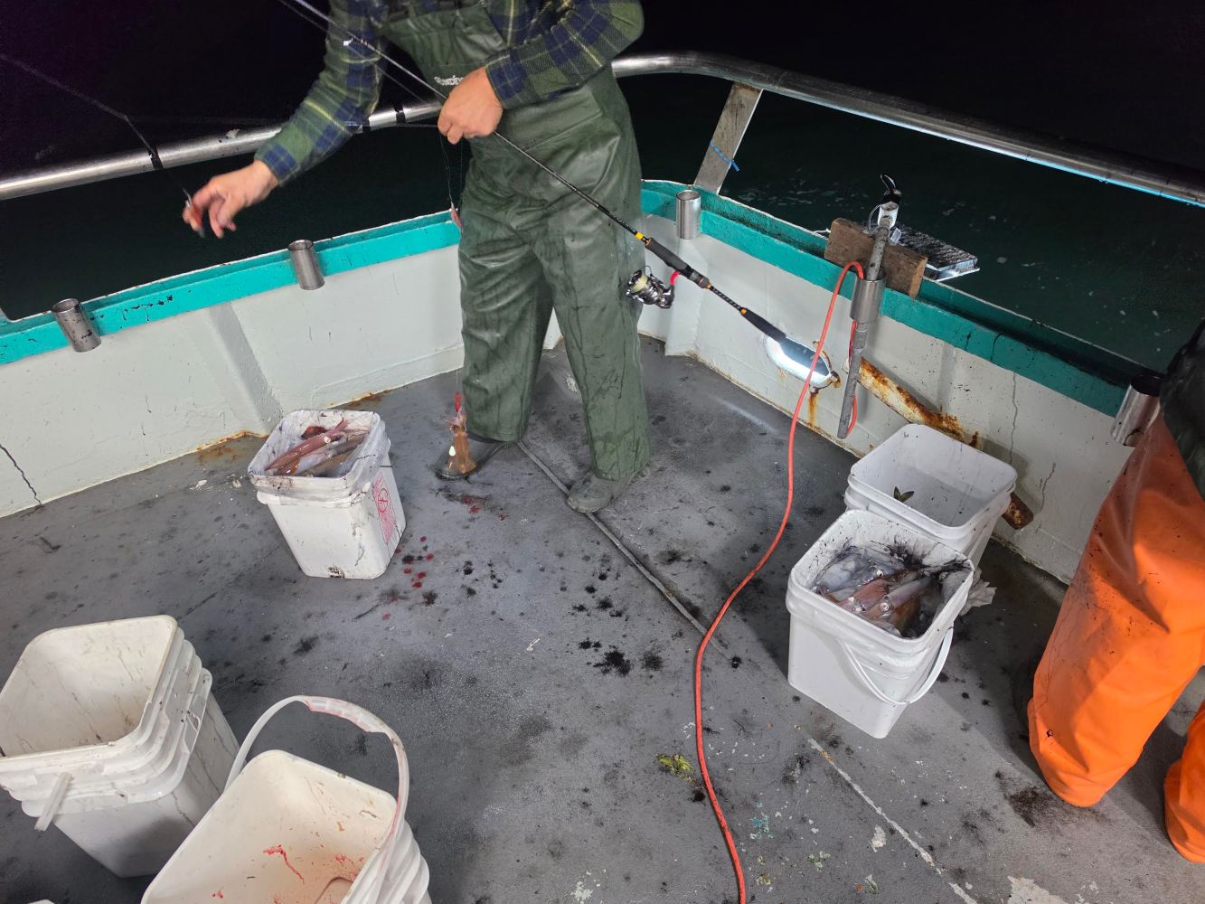 Person fishing at night on a boat with buckets and fishing gear.