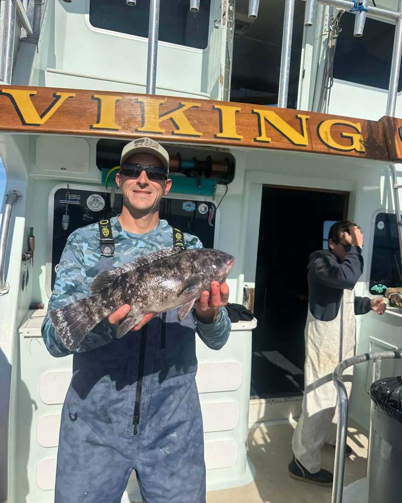 Man on boat holding a large fish, sign above reads 'VIKING'; another person in background adjusting cap.