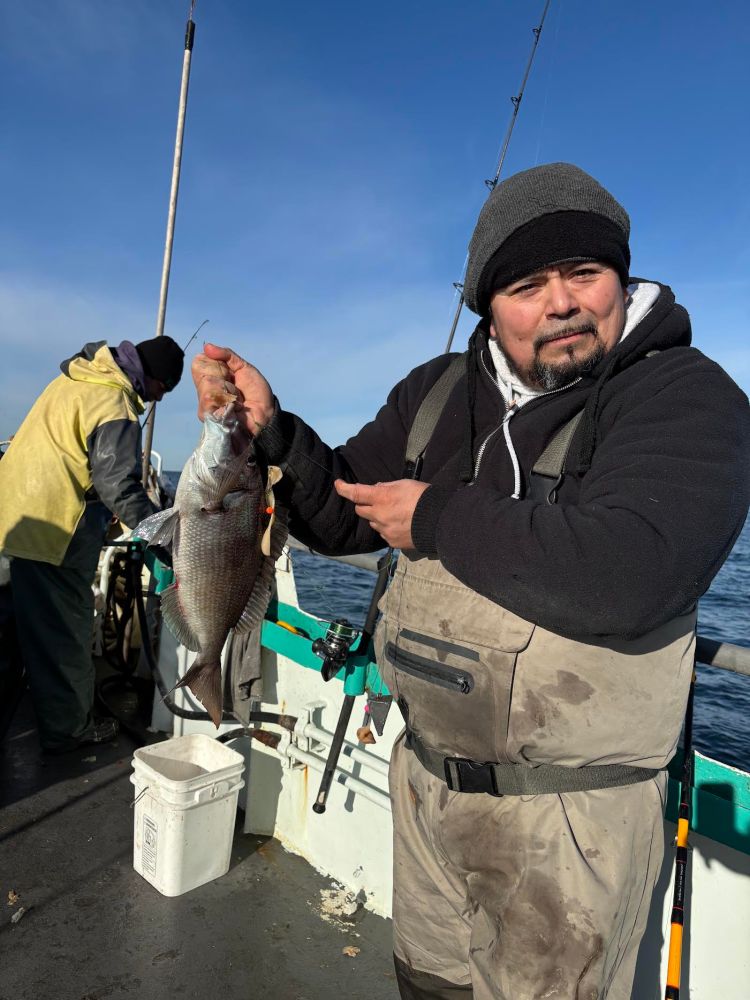 Man on boat holding a large fish, wearing waders and a beanie.