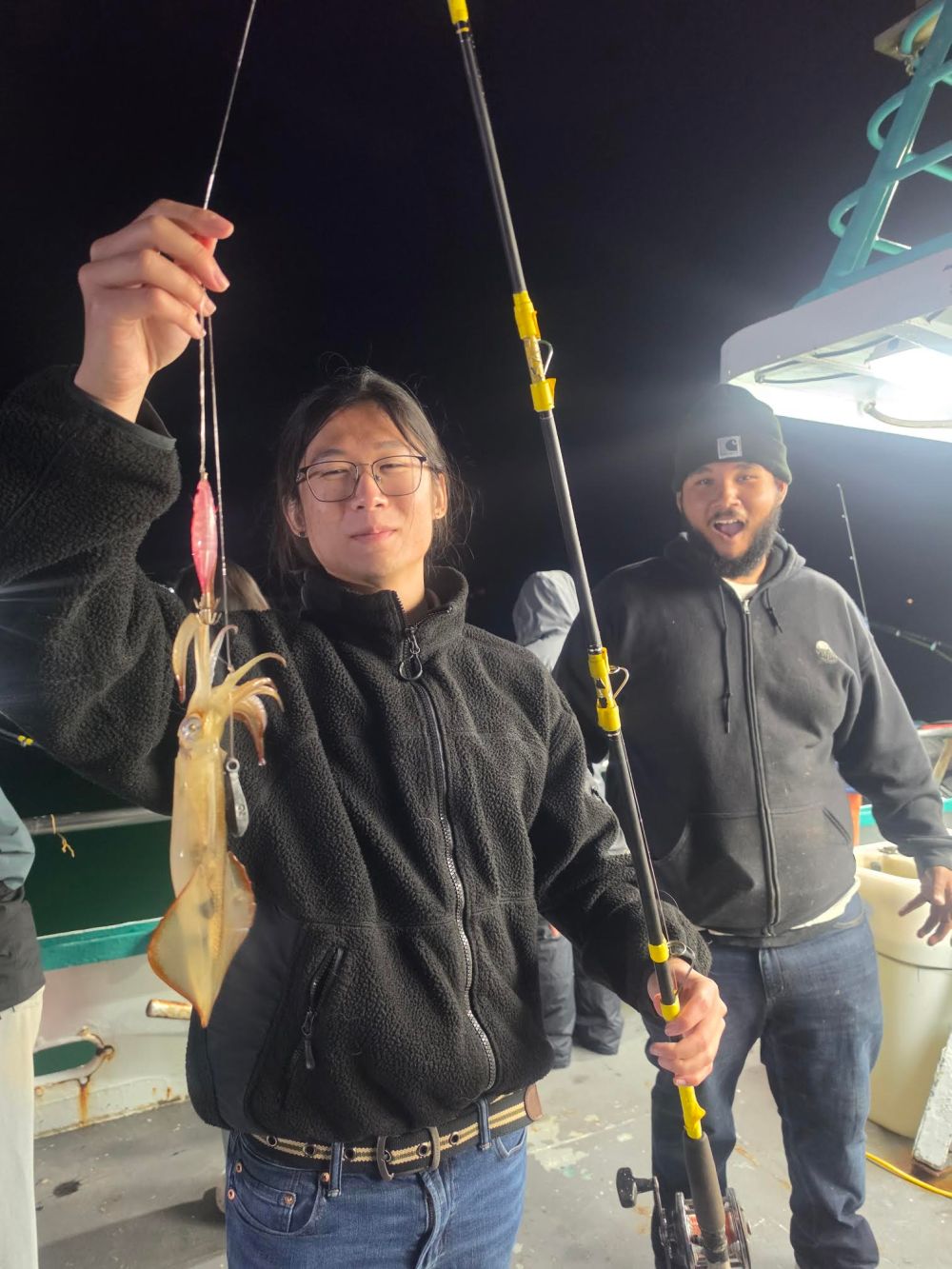 Person holding a fishing rod with a squid caught, standing next to another person on a boat at night.