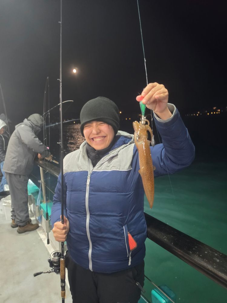 Person on a boat at night holding a caught squid and fishing rod, smiling.