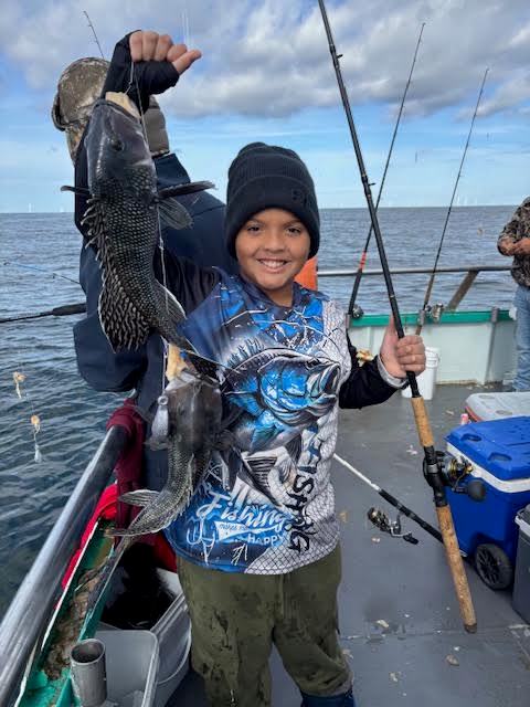 Child holding a fish on a boat, wearing a beanie and fishing gear, with the ocean in the background.