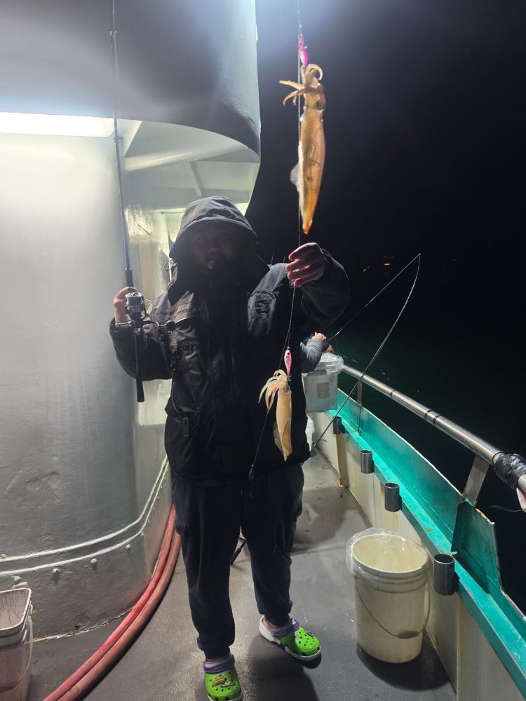 Person in a hooded jacket on a boat holding a fishing line with two caught squid at night.