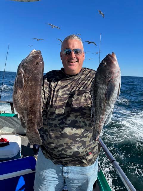 Person in camo holding two large fish on a boat with seagulls and ocean in background.