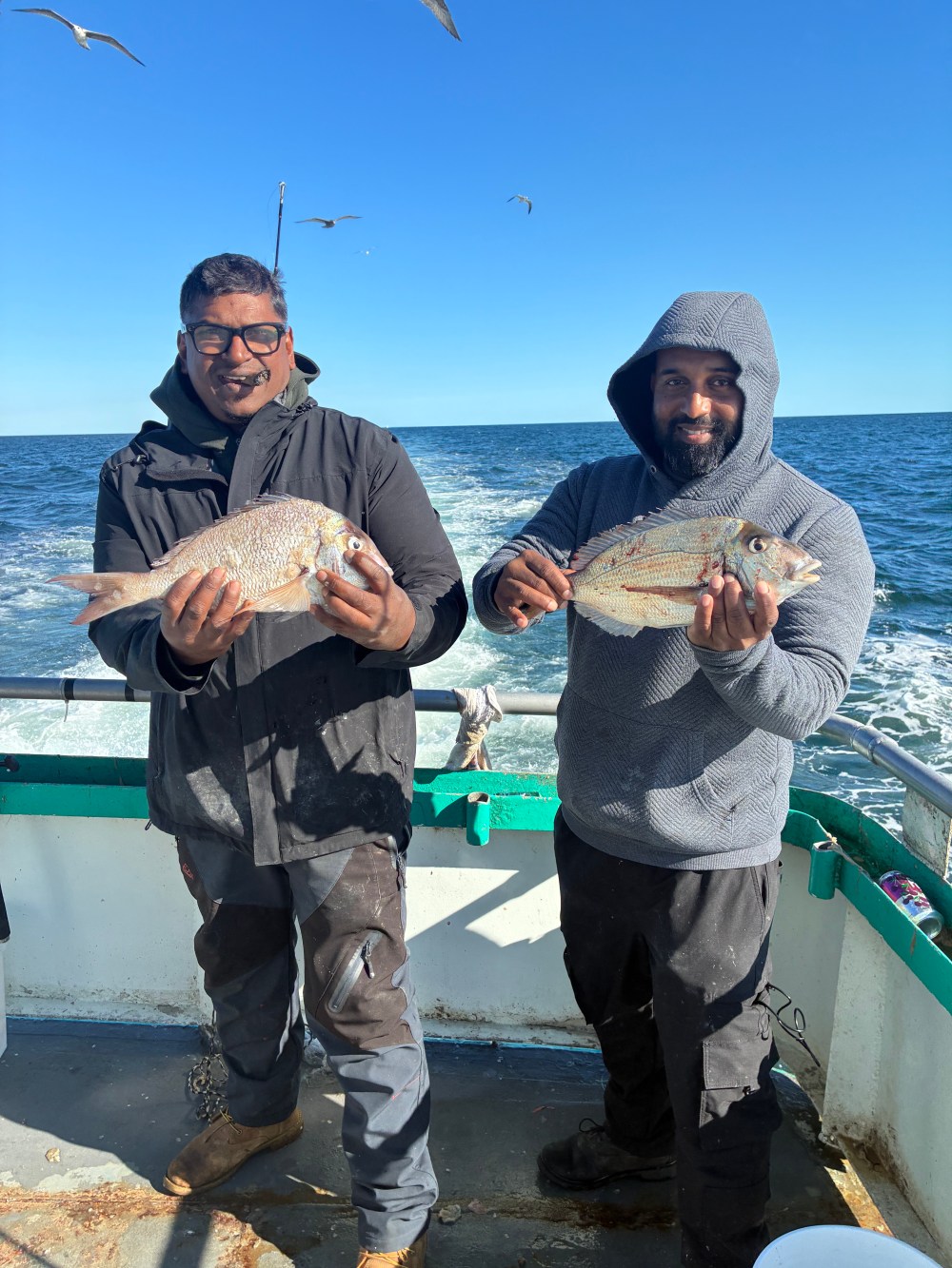 Two smiling men on a boat holding large fish with ocean backdrop.