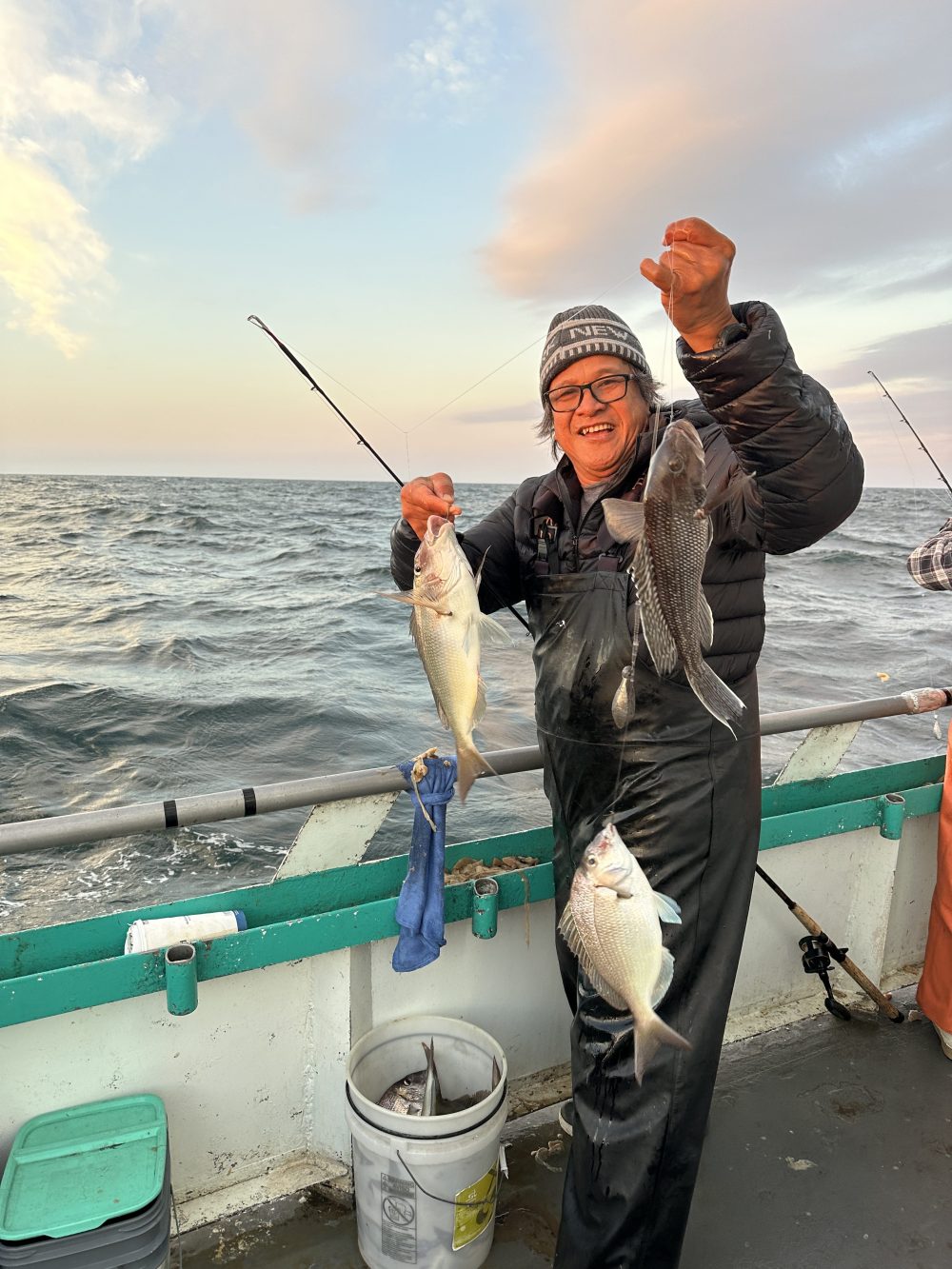 Person holding several fish on a boat at sea, smiling and wearing a beanie.