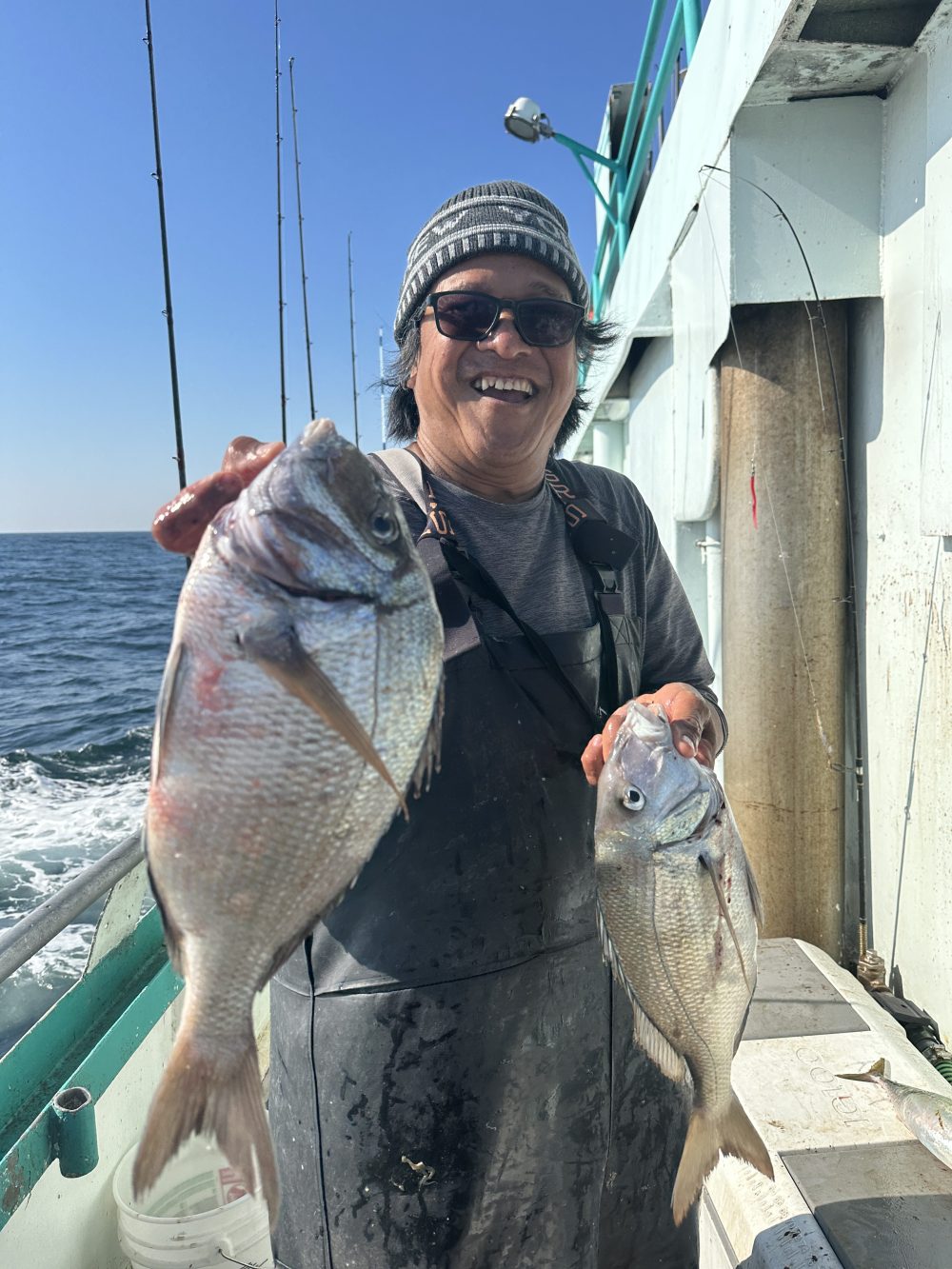 Person holding two fish on a boat, smiling, with fishing rods in the background.