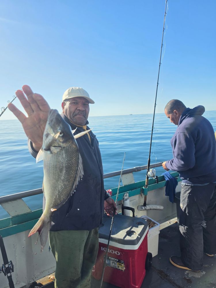 Two people fishing on a boat, one holding a fish, clear sky and sea.
