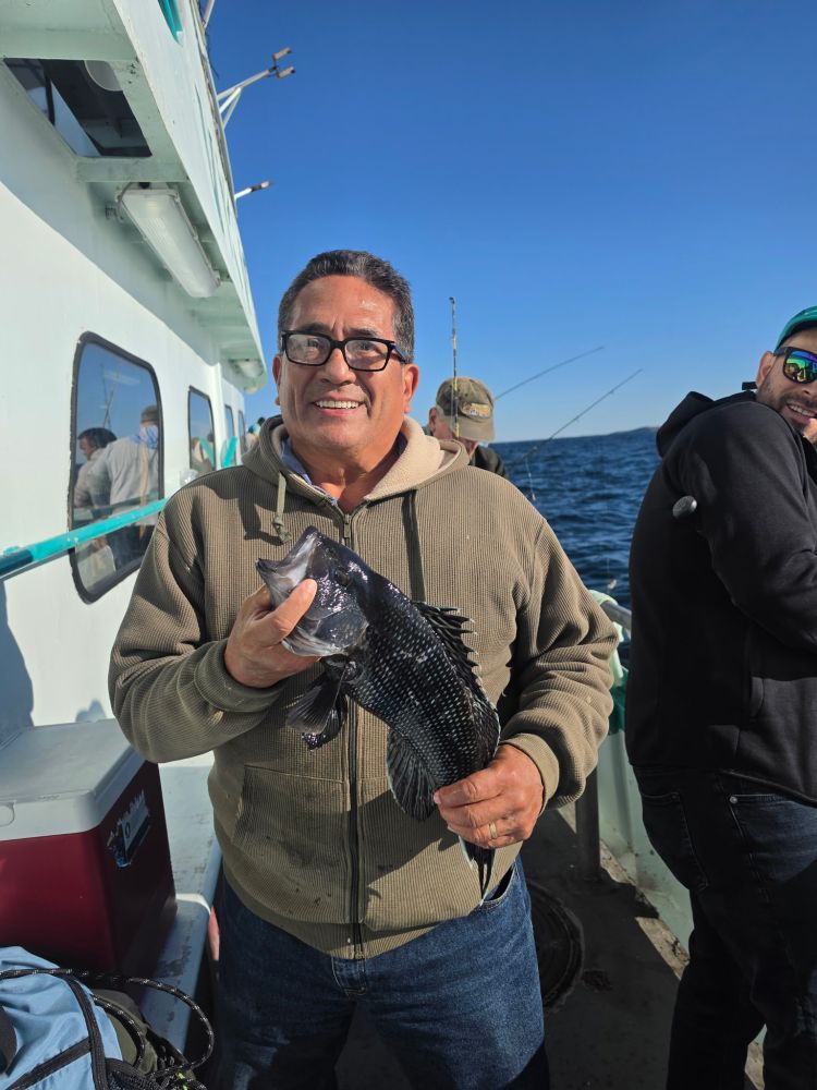 Man holding a fish on a boat with a smiling expression, under clear blue skies.